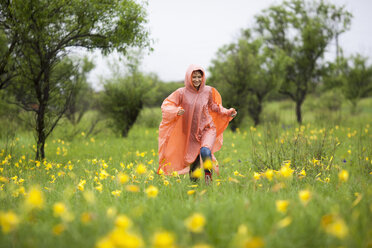 Woman wearing raincoat running amidst yellow flowering plants in rainy season - FSIF01818