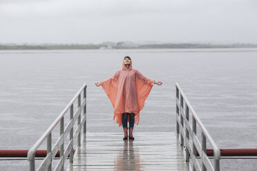 Woman wearing raincoat enjoying rainy season while standing on jetty - FSIF01819