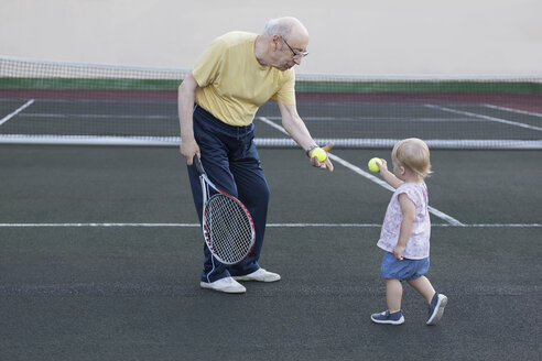 Girl giving tennis ball to grandfather while standing at playing field - FSIF01830
