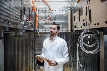 Man wearing lab coat and safety goggles at machine holding tablet - DIGF03393