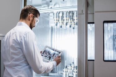 Man wearing lab coat and safety goggles at machine in factory looking at tablet - DIGF03398