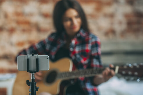 Smart Phone in Einbeinstativ mit Frau spielt Gitarre im Hintergrund zu Hause - FSIF01843