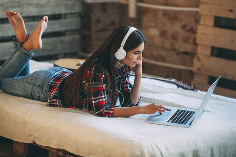 Junge Frau hört Musik und benutzt einen Laptop auf dem Bett zu Hause, lizenzfreies Stockfoto