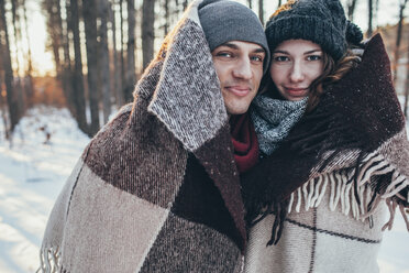 Porträt eines lächelnden Paares, das in eine Decke eingewickelt auf einem schneebedeckten Feld steht - FSIF01849