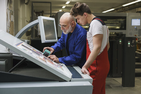 Ein älterer Arbeiter zeigt einem Kollegen die Qualität des Ausdrucks mit einem Scanner an einer Druckmaschine - FSIF01915