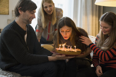 Familie betrachtet junge Frau beim Ausblasen von Geburtstagskerzen zu Hause - FSIF01928