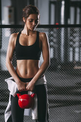 Frau hebt Kettlebell, während sie gegen den Zaun im Fitnessstudio steht - FSIF01941