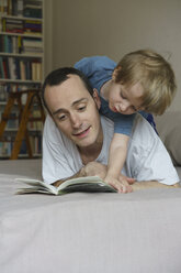 Father and son reading book while lying on bed at home - FSIF01954