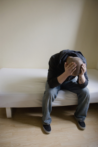 Junger Mann sitzt auf dem Bett und hält seinen Kopf in den Händen, lizenzfreies Stockfoto