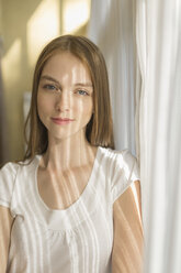 Portrait of smiling woman standing by curtains at home on sunny day - FSIF02173