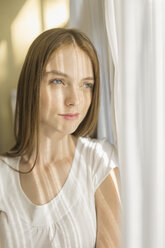 Thoughtful smiling woman standing by curtains at home on sunny day - FSIF02175