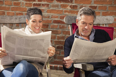 Happy couple reading newspaper against brick wall in back yard - FSIF02208