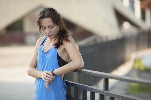 Schöne junge Frau schaut auf Fitness-Tracker, während sie an einem Geländer im Park steht - FSIF02238