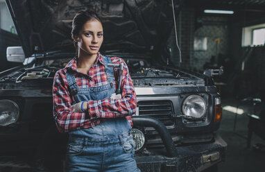 Porträt einer selbstbewussten Mechanikerin mit verschränkten Armen, die sich in einer Werkstatt an ein Auto lehnt - FSIF02273