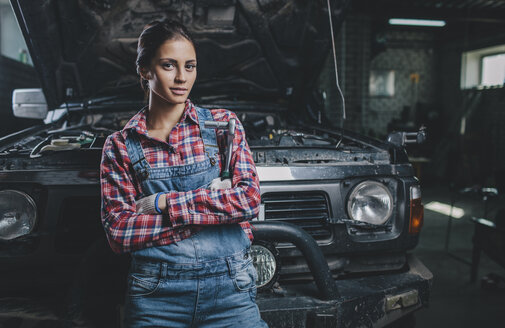 Porträt einer selbstbewussten Mechanikerin mit verschränkten Armen, die sich in einer Werkstatt an ein Auto lehnt - FSIF02273
