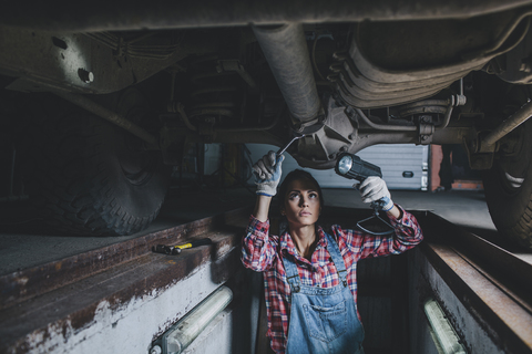 Mechanikerin mit Taschenlampe bei der Arbeit unter dem Auto in der Werkstatt, lizenzfreies Stockfoto