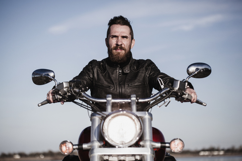 Porträt eines selbstbewussten Motorradfahrers auf einem Motorrad sitzend gegen den Himmel, lizenzfreies Stockfoto