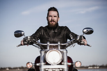 Porträt eines selbstbewussten Motorradfahrers auf einem Motorrad sitzend gegen den Himmel - FSIF02291