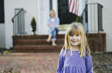 Junges Mädchen, das auf einem Weg vor einem Haus steht, während ihre Mutter auf der Türschwelle sitzt - FSIF02380