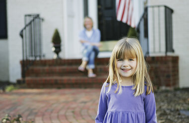 Young girl standing on a pathway in front of a house with her mother sitting behind on the doorstep - FSIF02380