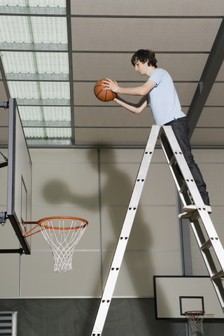 Ein junger Mann steht auf einer Leiter und bereitet sich darauf vor, einen Basketball in einen Basketballkorb zu werfen, lizenzfreies Stockfoto