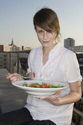 Eine junge Frau sitzt auf einer Dachterrasse und hält einen Teller Salat in der Hand - FSIF02525