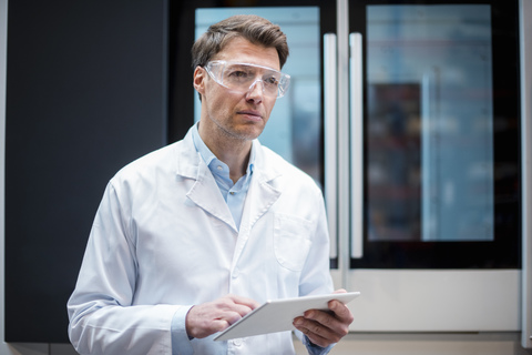 Porträt eines Mannes mit Laborkittel und Schutzbrille, der ein Tablet an einer Maschine hält, lizenzfreies Stockfoto