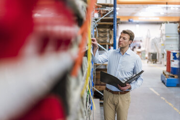 Man in factory storeroom holding folder checking shelf - DIGF03425