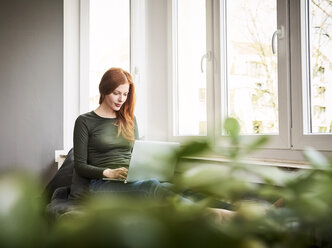 Redheaded woman sitting beside window using laptop - FMKF04858