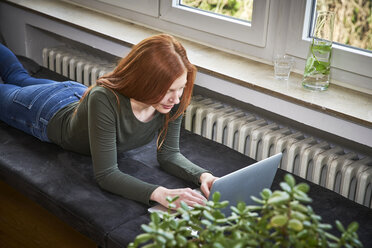 Rothaarige Frau liegt auf einer Bank in der Nähe der Heizung und benutzt einen Laptop - FMKF04860