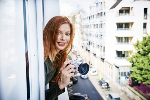 Porträt einer lächelnden rothaarigen Frau mit Kamera, die sich aus dem Fenster lehnt - FMKF04865