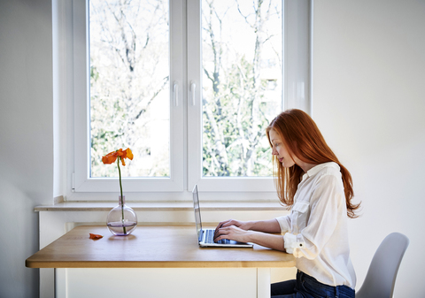 Redheaded woman sitting at table in front of window using laptop stock photo