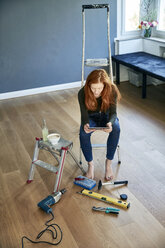 Redheaded woman sitting on step ladder using tablet - FMKF04876