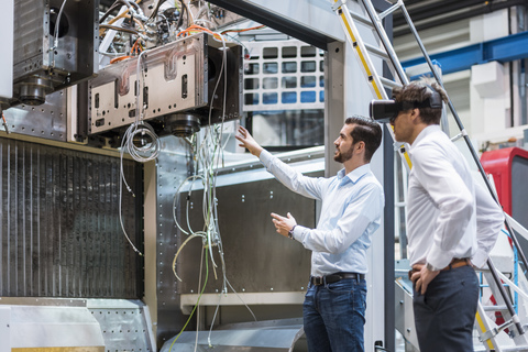 Man explaining machine to colleague wearing VR glasses in factory stock photo