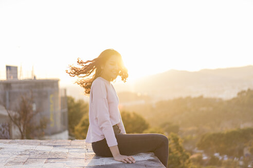 Junge Frau sitzt bei Sonnenuntergang auf einer Mauer und wirft ihr Haar - AFVF00118