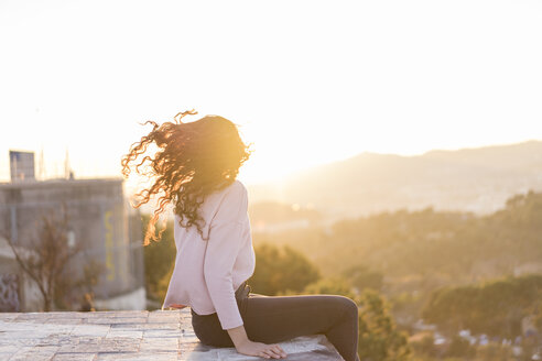 Junge Frau sitzt bei Sonnenuntergang auf einer Mauer und wirft ihr Haar - AFVF00119