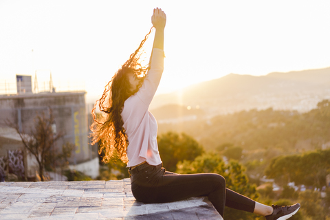 Junge Frau sitzt bei Sonnenuntergang auf einer Mauer und zieht an ihren Haaren, lizenzfreies Stockfoto