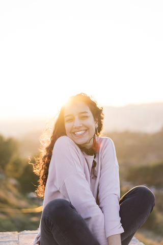 Portrait of happy young woman with long hair sitting on a wall at sunset stock photo