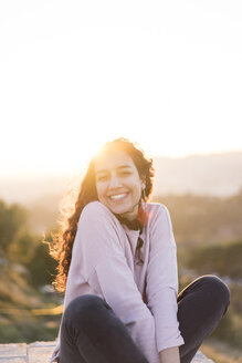 Porträt einer glücklichen jungen Frau mit langen Haaren, die bei Sonnenuntergang an einer Mauer sitzt - AFVF00125