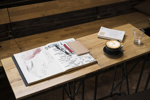 Tisch in einem Café mit Kaffeebecher, Notizbüchern, Stiften und einem Glas Wasser - SBOF01364