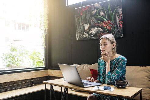 Junge Frau mit grünem Kleid sitzt in einem Café und arbeitet an ihrem Laptop - SBOF01366