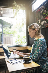 Junge Frau mit grünem Kleid sitzt in einem Café und arbeitet an ihrem Laptop - SBOF01368