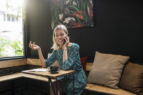 Junge lächelnde Frau mit grünem Kleid sitzt in einem Café und spricht mit jemandem über ihr Smartphone - SBOF01369