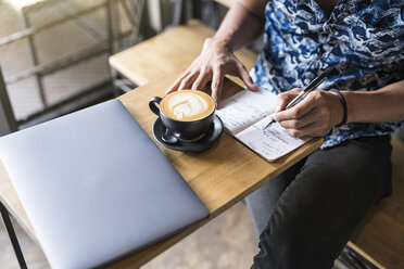 Künstlerin schreibt in ein Notizbuch in einem Café mit Kaffee und Laptop auf dem Tisch - SBOF01388