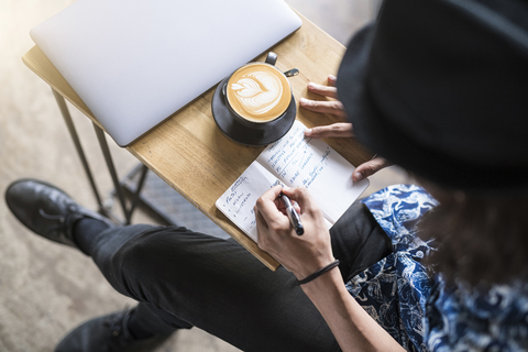 Künstlerin schreibt in ein Notizbuch in einem Café mit Kaffee und Laptop auf dem Tisch, lizenzfreies Stockfoto