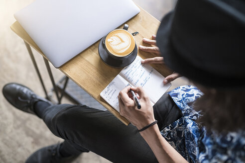 Künstlerin schreibt in ein Notizbuch in einem Café mit Kaffee und Laptop auf dem Tisch - SBOF01389