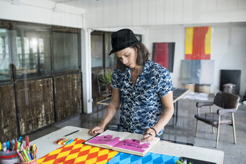 Künstler bei der Arbeit, der in seinem Loft-Atelier in ein Notizbuch zeichnet, mit Gemälden und Kunstwerken im Hintergrund - SBOF01402