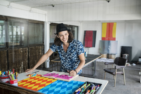 Lächelnder Künstler bei der Arbeit, der in seinem Loft-Atelier mit Gemälden und Kunstwerken im Hintergrund in ein Notizbuch zeichnet, lizenzfreies Stockfoto