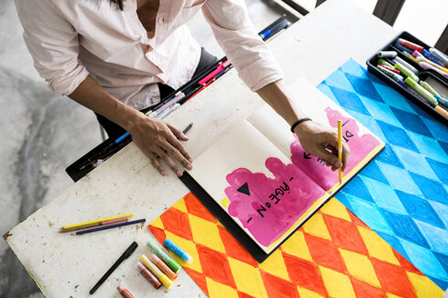 Hände eines Künstlers, der in ein Notizbuch zeichnet, während sein Kunstwerk auf dem Tisch liegt - SBOF01407