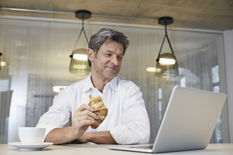 Geschäftsmann benutzt Laptop und isst Croissant, lizenzfreies Stockfoto